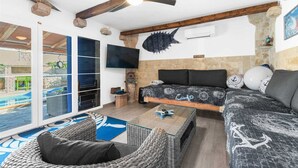 Amplia sala de estar con una cama grande, un sofá y una mesa de comedor. Hay un balcón con vistas a una piscina y un jardín.