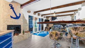 Amplia sala de estar con chimenea de piedra, vigas de madera y una gran decoración con un ancla. Hay dos taburetes de bar con tapicería floral y un tapete azul estampado en el suelo.