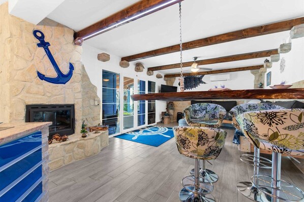 A spacious living area with a stone fireplace, wooden beams, and a large anchor decoration. There are two bar stools with floral upholstery and a blue patterned rug on the floor.
