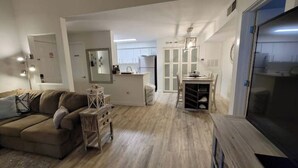 A modern living room with a sofa, a small table, and a TV. There is a kitchen area with cabinets and a refrigerator in the background.