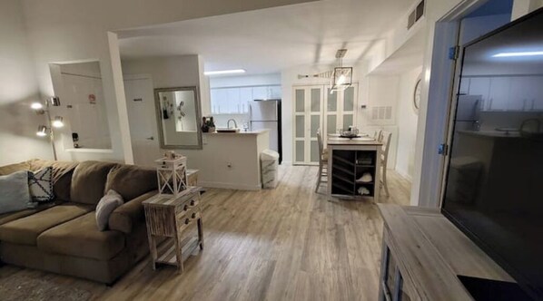 A modern living room with a sofa, a small table, and a TV. There is a kitchen area with cabinets and a refrigerator in the background.