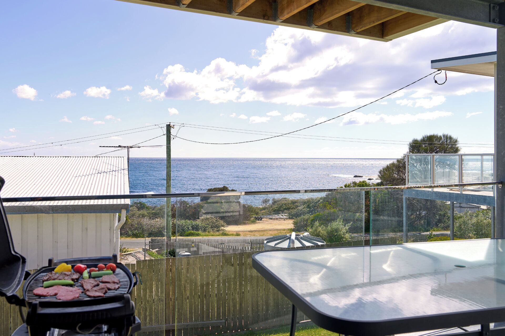Un balcon avec vue sur la mer, un barbecue avec de la viande et une table en verre.