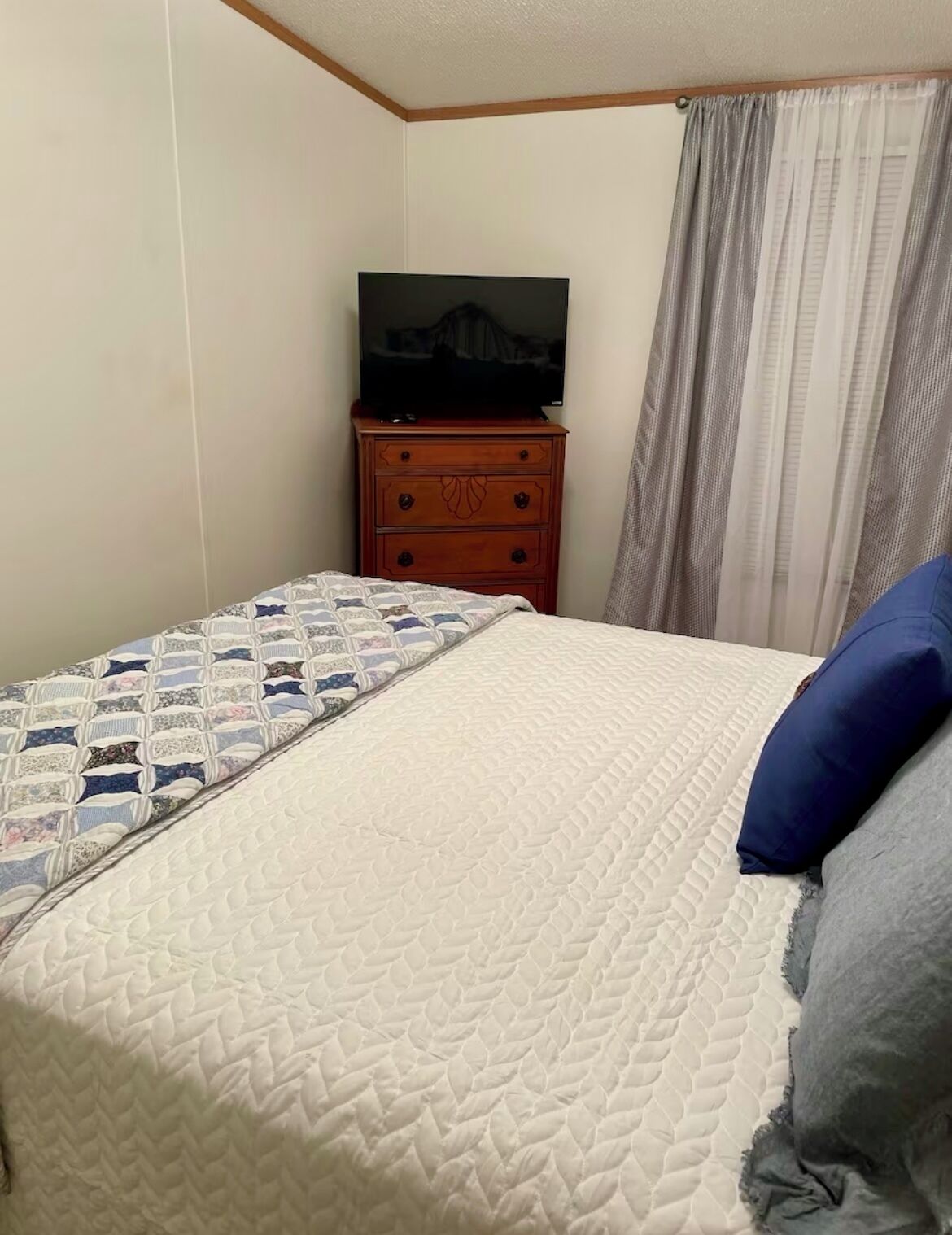 Un dormitorio con cama, cómoda de madera, televisor y ventana con cortinas.