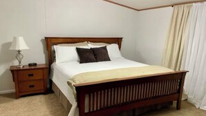 Un dormitorio con una cama de madera, ropa de cama blanca, una mesita de noche con una lámpara y una alfombra color beige.