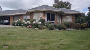 A two-story house with a garage, a covered porch, and a well-maintained front yard with various plants and flowers. - Cherry Tree Lane Accommodation, Quiet location bordered by farmland. (Te Awamutu)