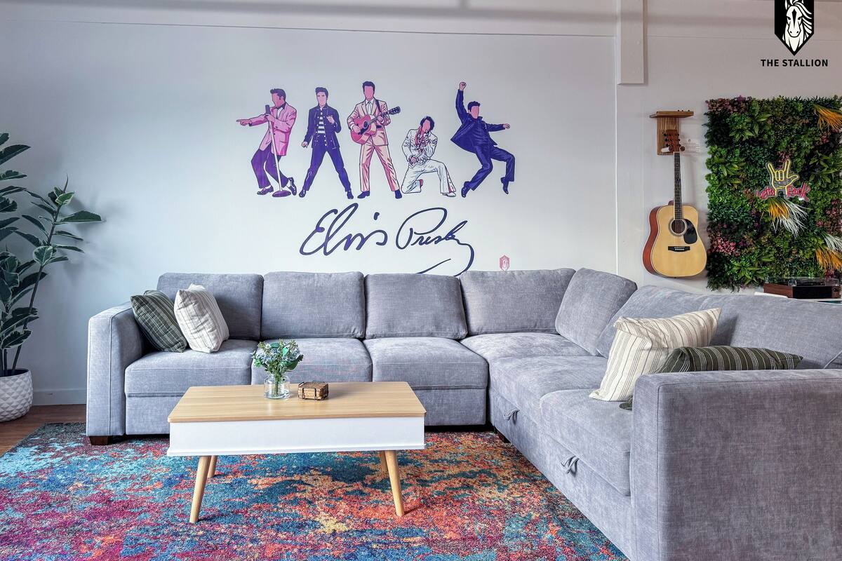 A living room with a grey sectional sofa, a colorful rug, a white coffee table, and a wall mural of Elvis Presley.