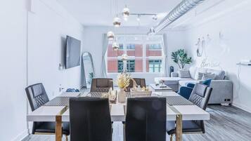 A modern living room with a sofa, a dining table with chairs, a TV mounted on the wall, and a large window with a view of buildings.
