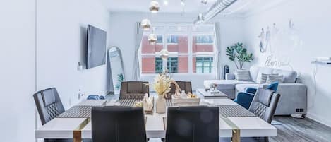 Een moderne woonkamer met een bank, een eettafel met stoelen, een aan de muur bevestigde televisie en een groot raam met uitzicht op gebouwen.