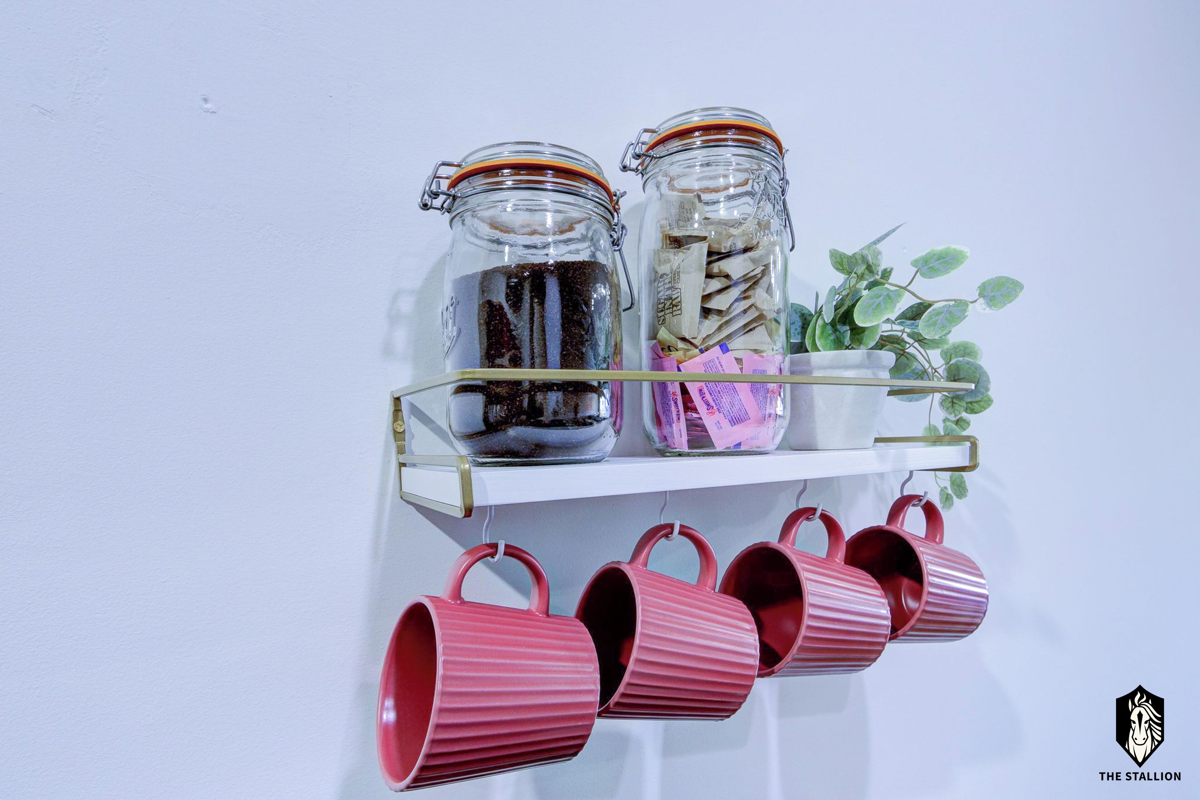 Dos frascos de vidrio sobre un estante blanco, con una planta y tazas rosas debajo.