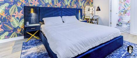 A modern bedroom with a large bed, a vibrant floral wall, a blue headboard, and a nightstand with a lamp.