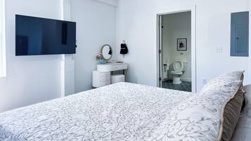 A bedroom with a bed, a TV mounted on the wall, a dresser, and a bathroom visible through an open door.
