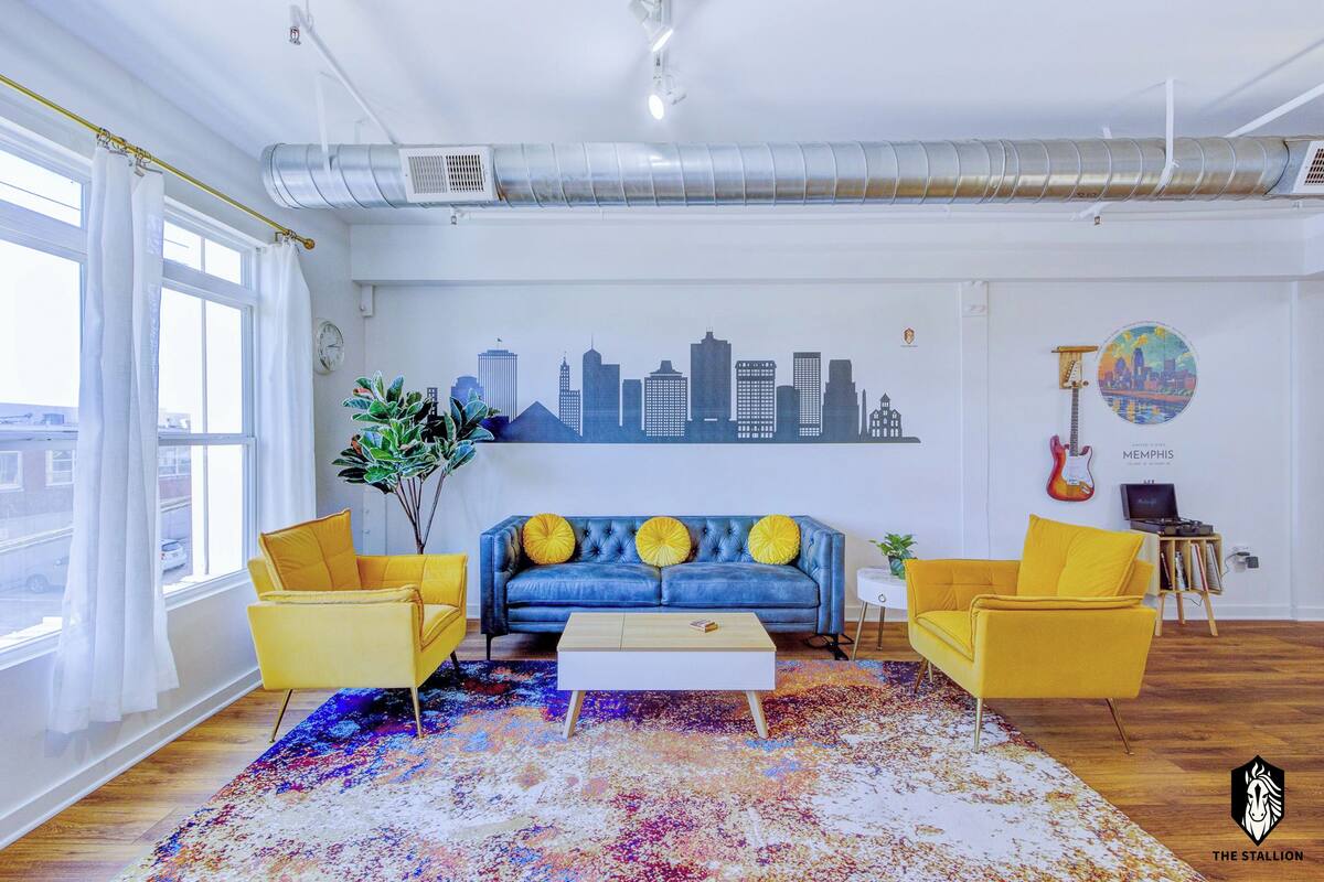 A modern living room with a blue sofa, yellow chairs, a coffee table, and a cityscape mural on the wall.