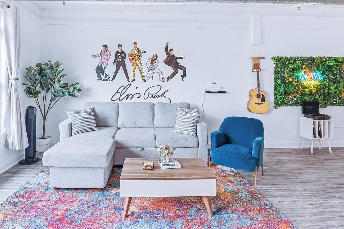 A modern living room with a grey sofa, a blue armchair, a coffee table, and a colorful rug. There's a wall art depicting musicians and a sign that says 'Elvis's Place'.