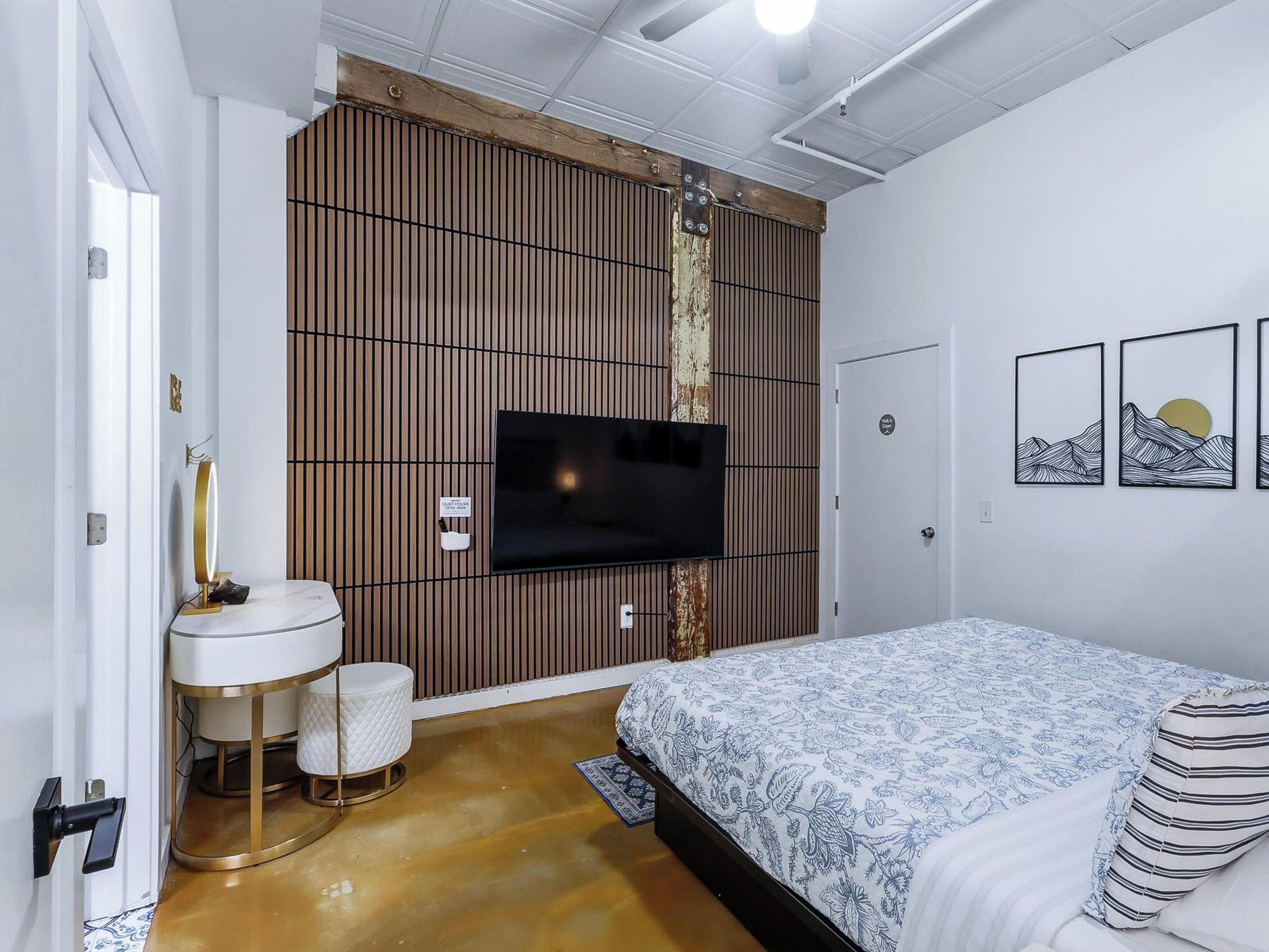 Un dormitorio con cama, televisor montado en una pared de paneles de madera, mesita de noche y un pequeño espejo redondo.