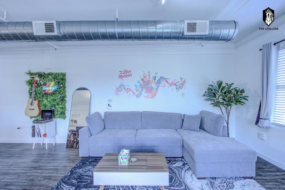 A modern living room with a grey sofa, a coffee table, and a decorative wall with a guitar and greenery.