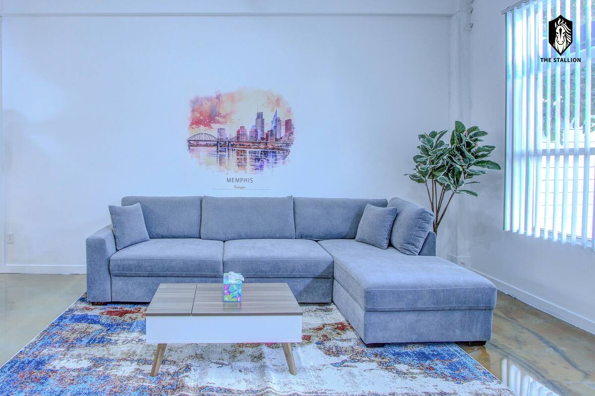 A modern living room with a grey sectional sofa, a wooden coffee table, and a colorful rug. A cityscape poster is on the wall, and a plant is beside the window.