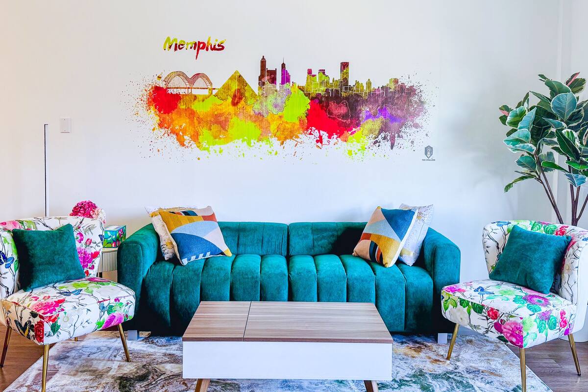 A teal sofa with floral-patterned armchairs, a wooden coffee table, and a vibrant cityscape mural on the wall.
