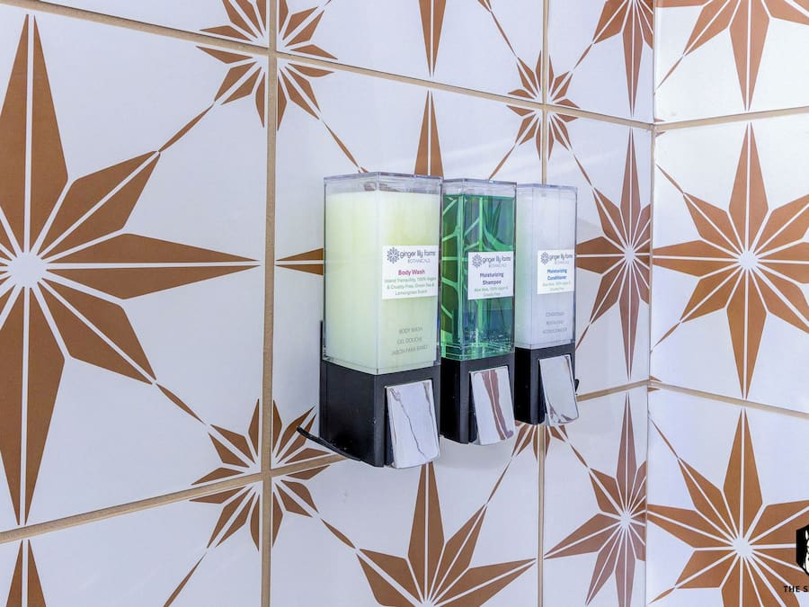 A wall-mounted dispenser with three bottles labeled for body wash, moisturizing shampoo, and conditioning cream.
