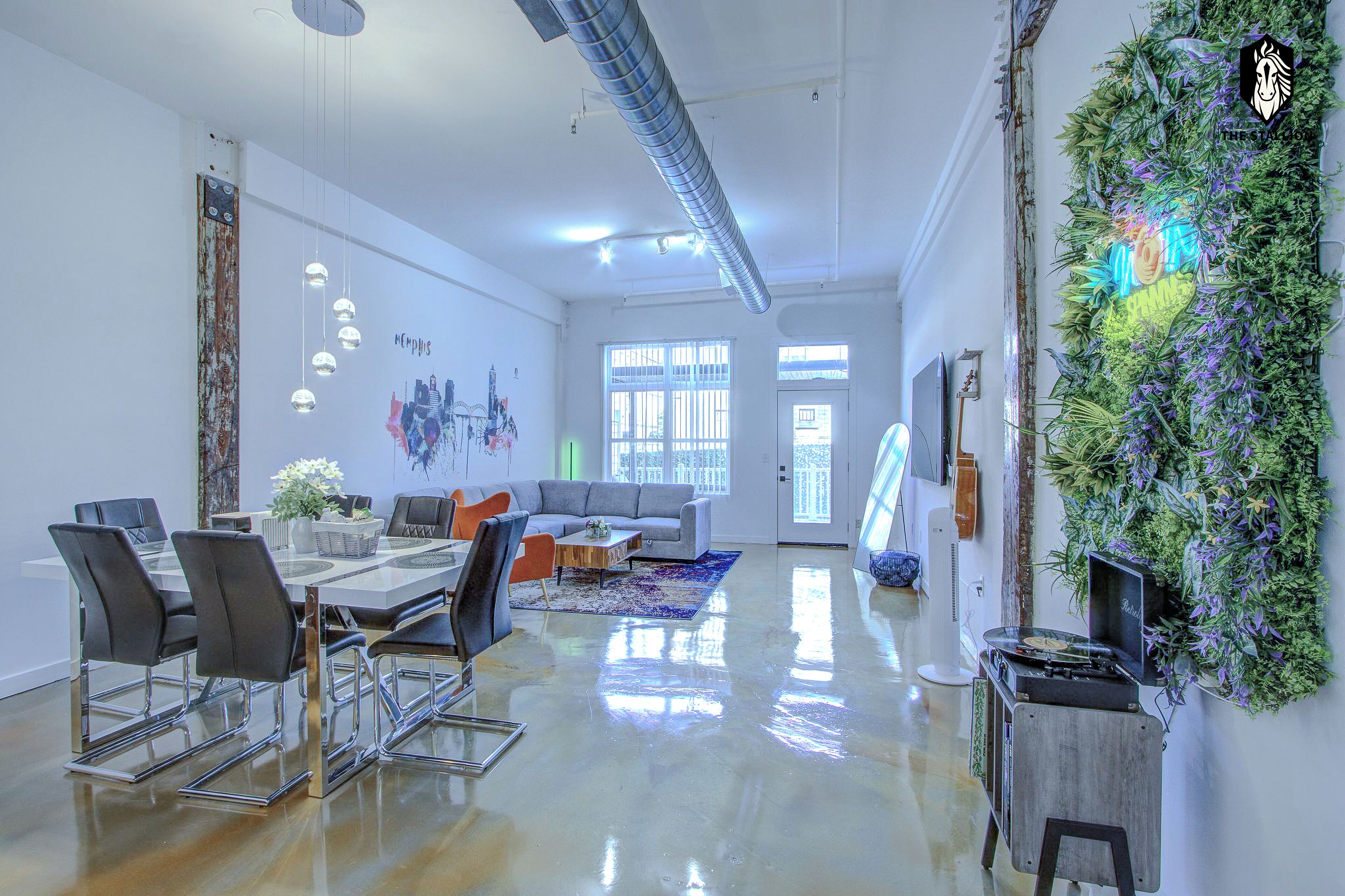 Un espacio de oficina moderno con una mesa de cristal, sillas, un sofá y una pared decorativa con plantas.