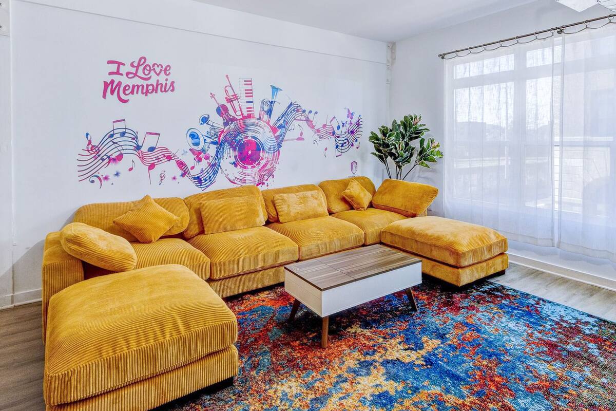 A modern living room with a yellow sectional sofa, a coffee table, and a colorful abstract wall mural.