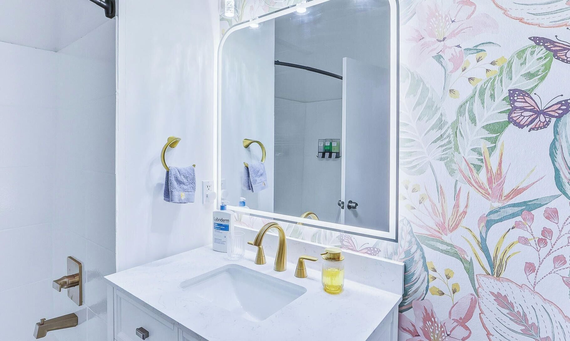 A bathroom with a white sink, gold faucets, and a mirror with a floral wallpaper.