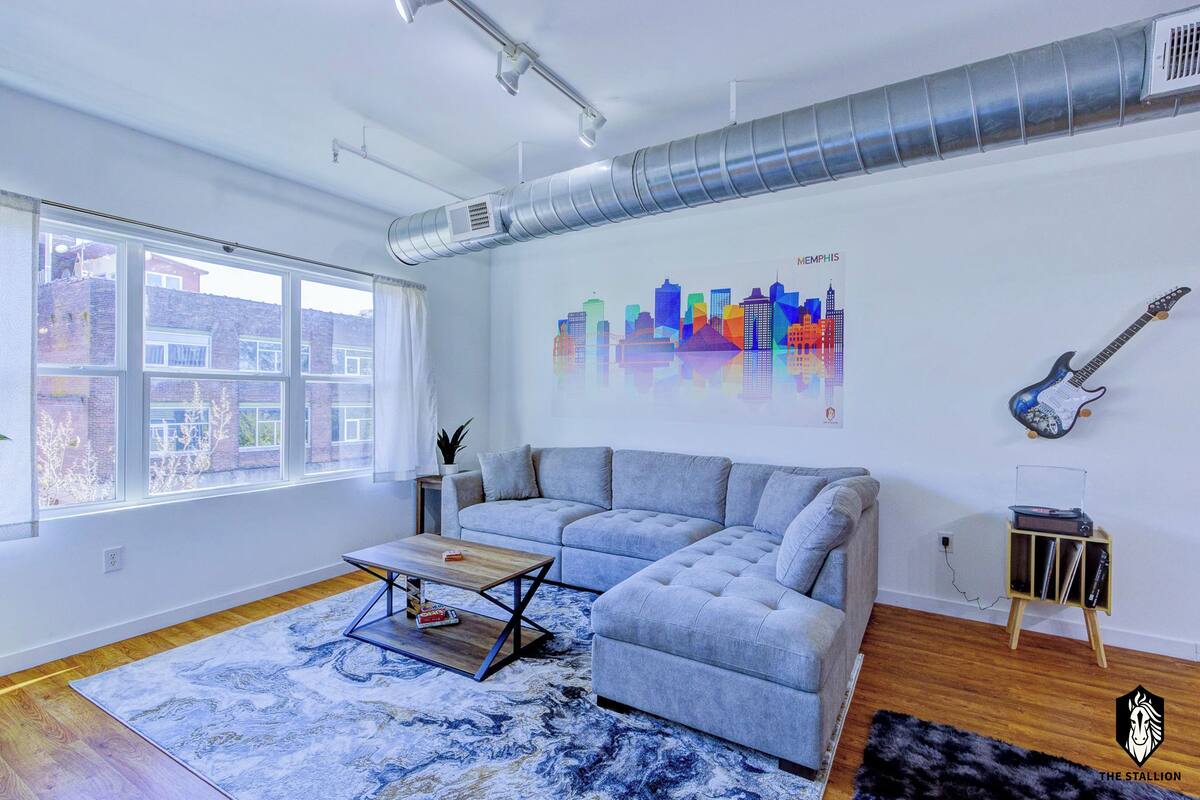 A modern living room with a grey sectional sofa, a coffee table, a rug, a city skyline mural, a guitar, and a small cabinet.