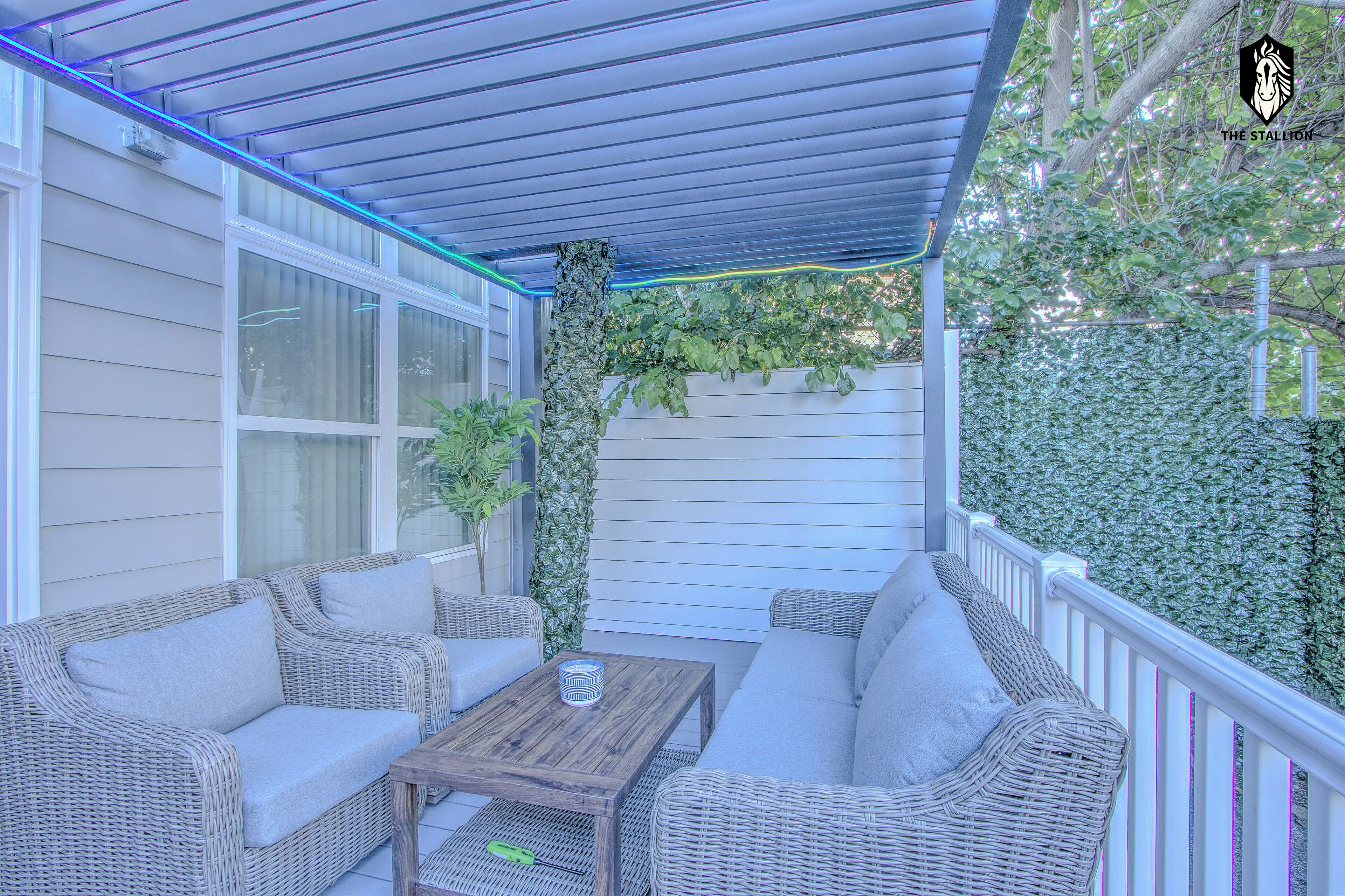 Eine überdachte Terrasse mit Korbmöbeln und einem Tisch, umgeben von weißen Geländern und viel Grün.