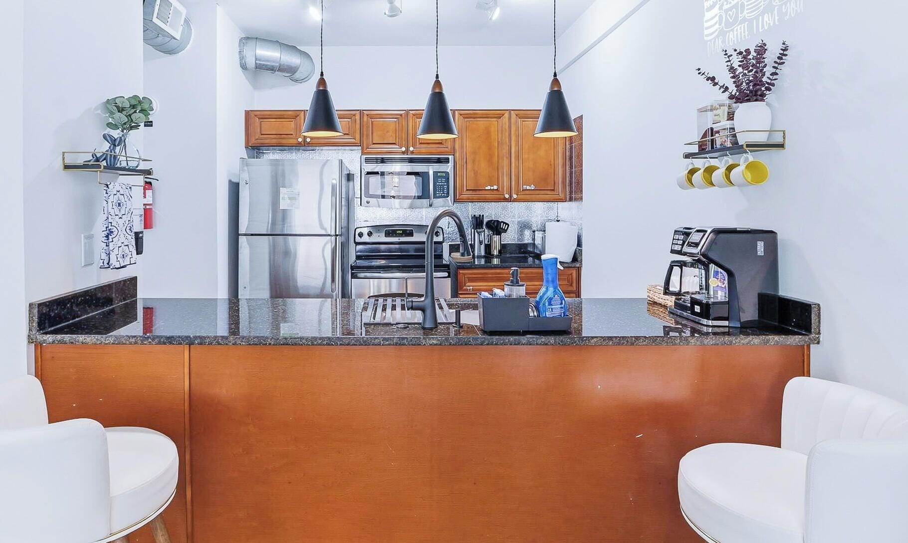 A modern kitchen with stainless steel appliances, wooden cabinets, a central island, pendant lighting, and a coffee machine.