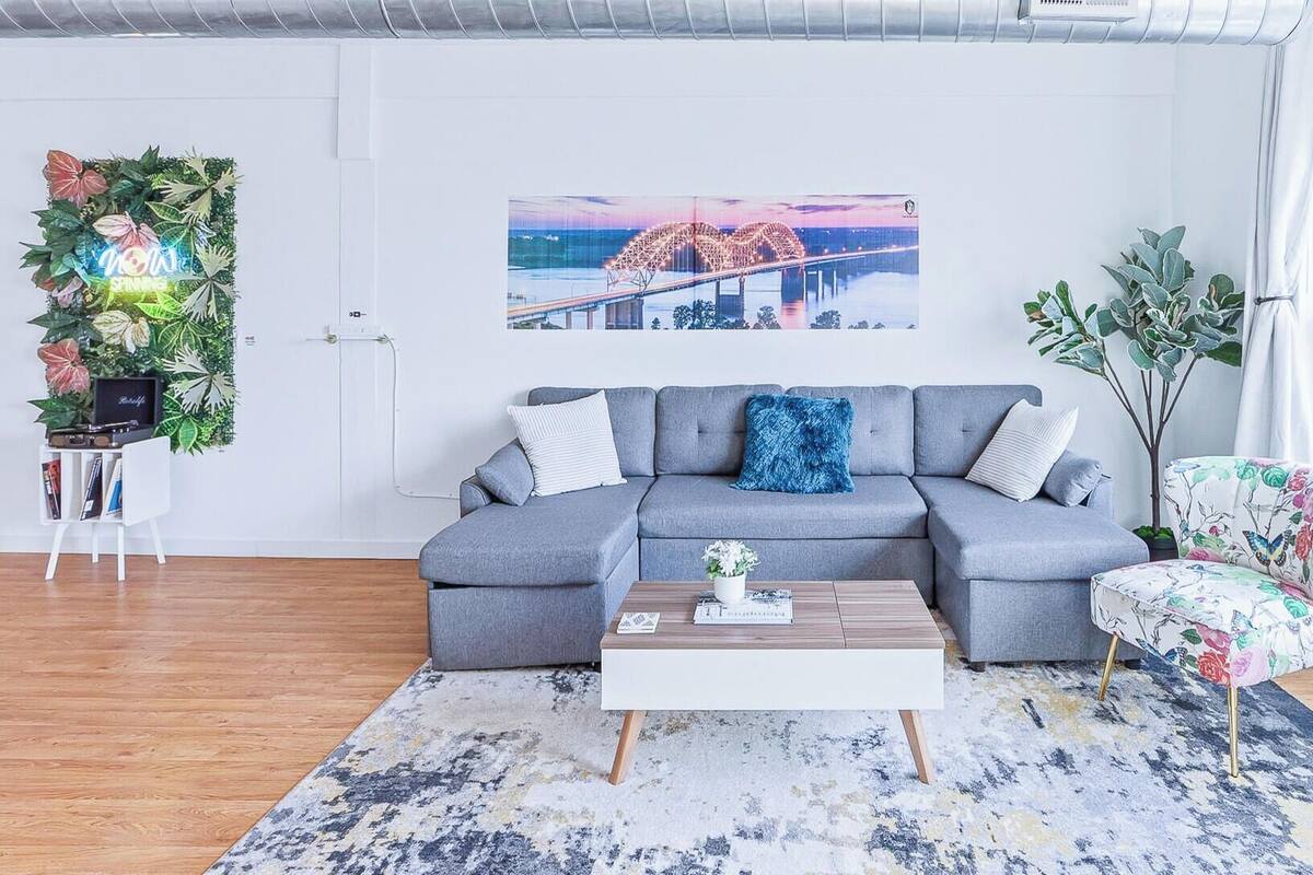 A modern living room with a grey sectional sofa, a coffee table, a floral armchair, a cityscape painting, and a green wall decoration.