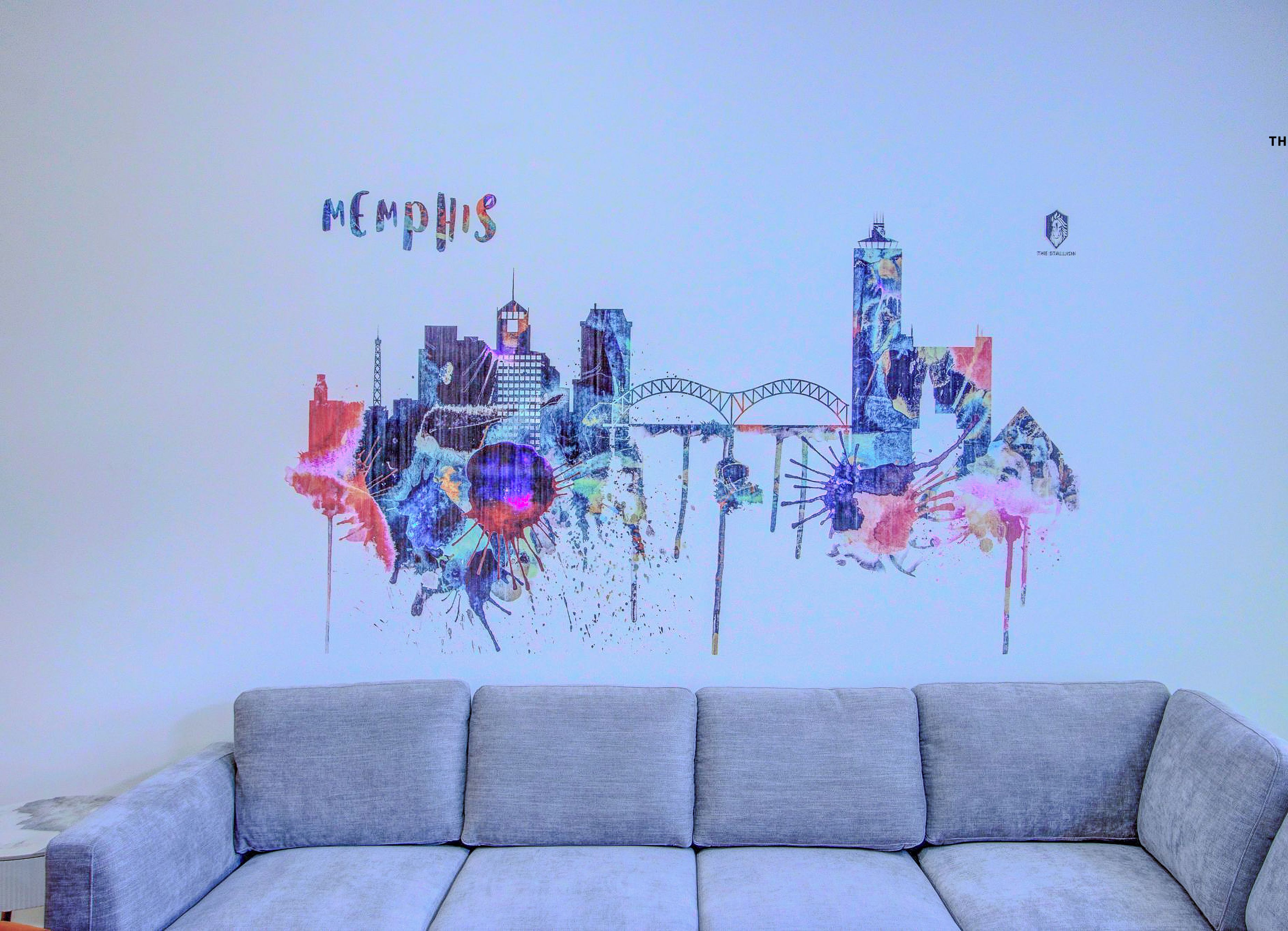 Decoración mural de un paisaje urbano colorido con la palabra "Memphis" en la parte superior, colocada sobre un sofá gris.