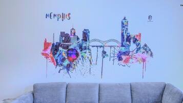 Decoración mural de un paisaje urbano colorido con la palabra "Memphis" en la parte superior, colocada sobre un sofá gris.