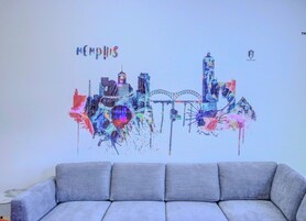 A colorful cityscape wall art with the word "Memphis" above it, placed above a grey sofa.