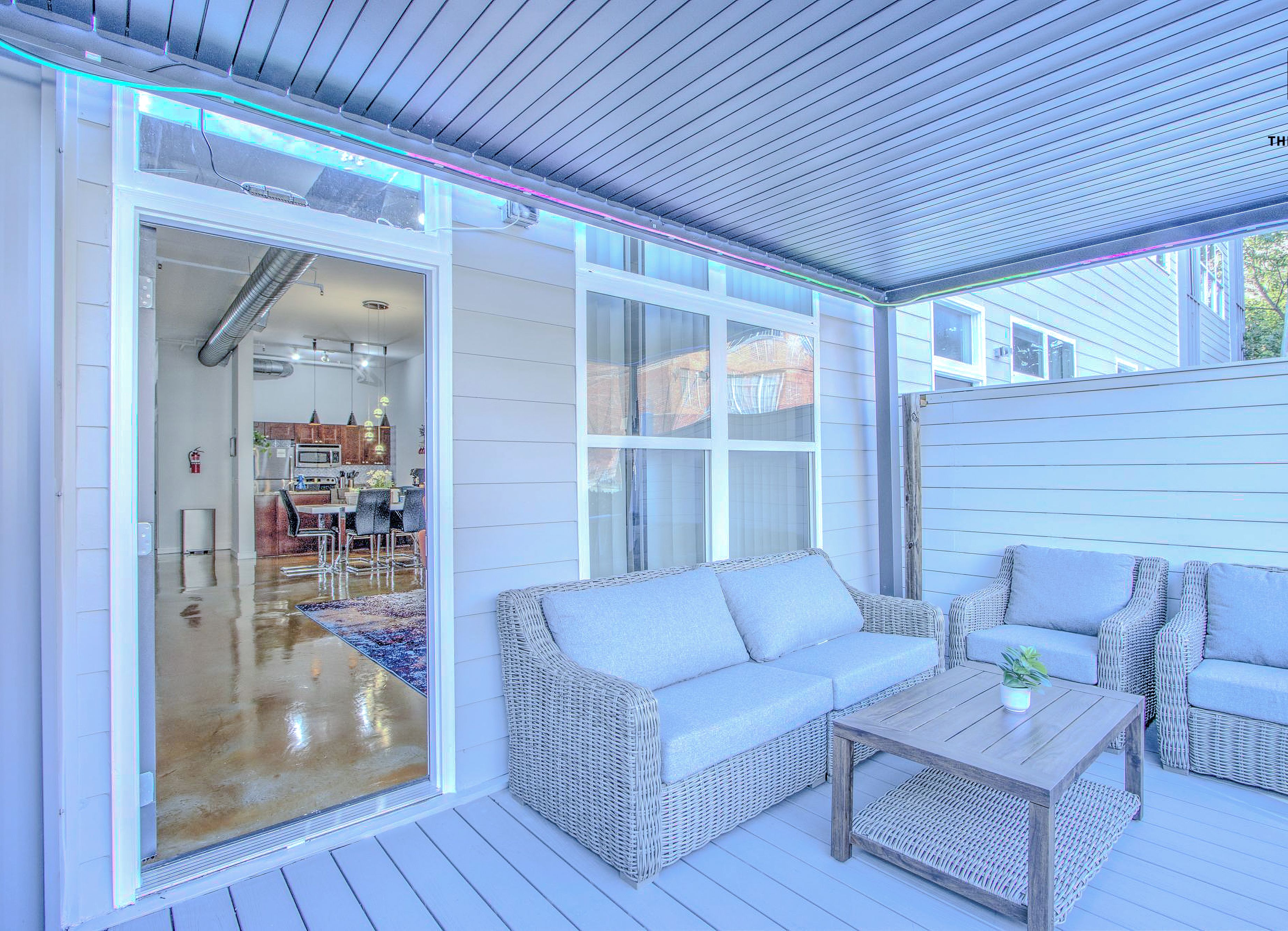 Un espacio exterior cubierto con mobiliario de mimbre, una mesa de madera y un ventanal corredero que da a un espacio interior.