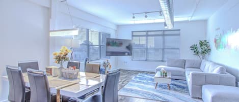 A modern living room with a glass dining table, grey sofas, and a large TV.