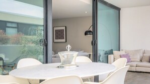 Een moderne buiten eethoek met een ronde tafel en stoelen, een tafel met glazen blad, een bank en een kunstwerk in een lijst aan de muur.