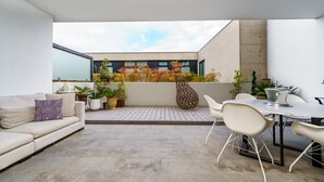 Een modern terras buiten met een bank, eettafel en stoelen, beplant met potten en uitzicht op een gebouw.
