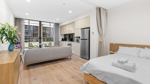 A modern hotel room with a bed, a bench, a kitchenette, and a view of the city through large windows.