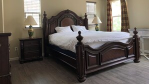 A bedroom with a large wooden bed, two bedside tables with lamps, a dresser, and a window with blinds.