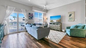 Un salon spacieux avec un canapé gris, des fauteuils turquoise et un plancher en bois. On y trouve deux ventilateurs de plafond, une grande fenêtre offrant une belle vue et un tableau accroché au mur.