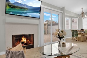 Een moderne woonkamer met een open haard, een glazen tafel en een televisie waarop een mooi landschap te zien is.