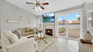 A modern living room with a white sofa, a glass coffee table, a flat-screen TV, a fireplace, and a view of a backyard through large sliding doors. - Henderson escape+ mini golf +peaceful stay (Henderson)
