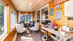 Ein gemütliches Blockhaus-Interieur mit Holzwänden und -decke, einem Essbereich mit Stühlen und einer Kaffeestation, sowie ein Ausblick durch große Fenster.