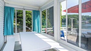 Eine überdachte Terrasse mit weißem Tisch und Bänken, große Fenster mit Vorhängen und Blick auf einen See und eine Anlegestelle.