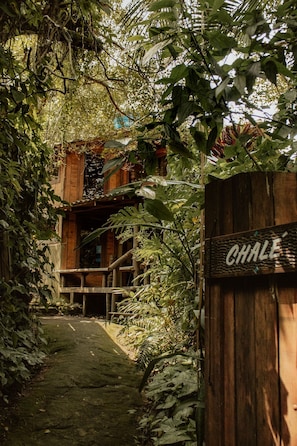 Une porte en bois, sur laquelle on peut lire l’inscription « CHALÉ », mène à un bâtiment rustique également en bois, entouré d’une végétation luxuriante.