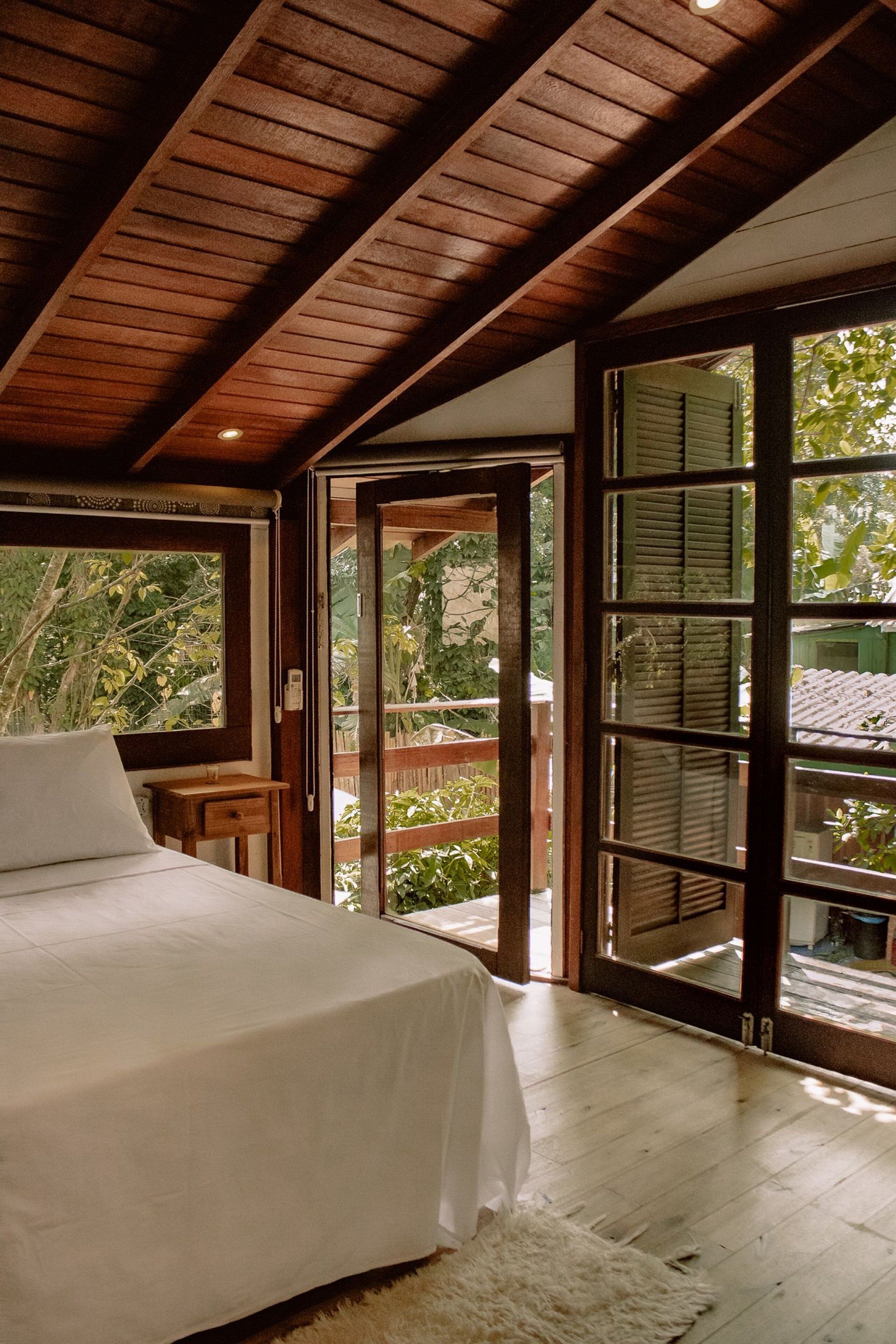 Une chambre dotée d’un grand lit, de poutres apparentes en bois au plafond, d’un plancher en bois et d’une porte-fenêtre coulissante donnant sur un espace extérieur.