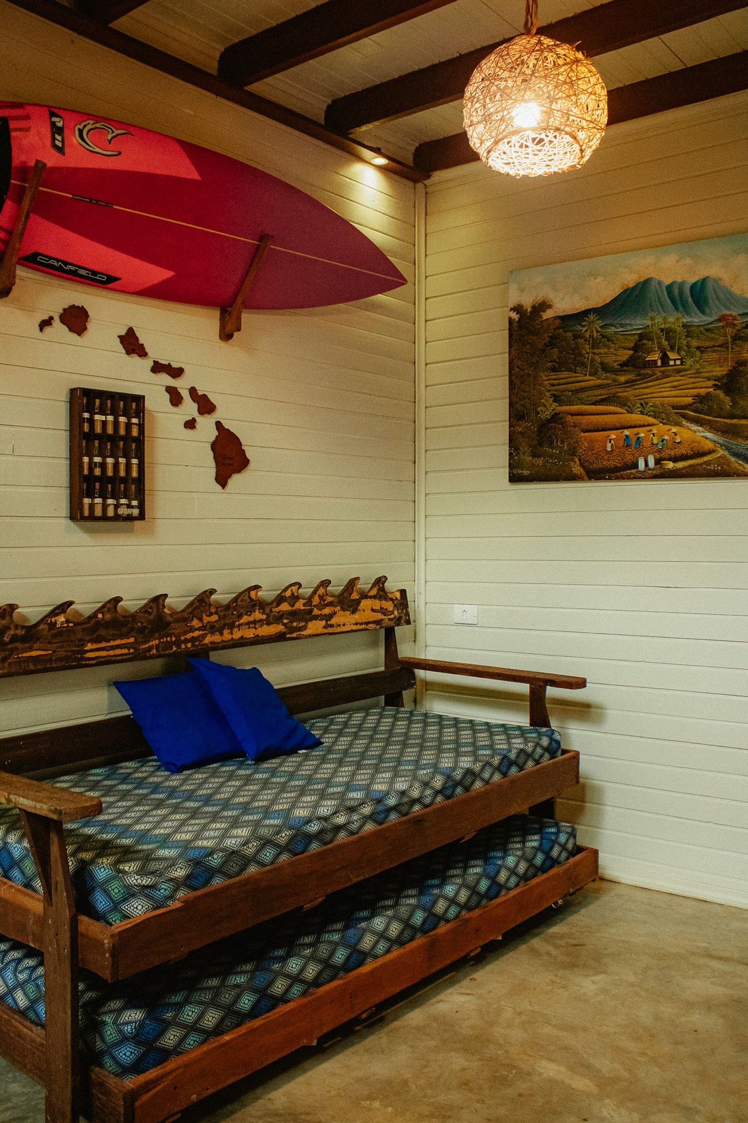 A room with a wooden bench bed, a red surfboard, a wooden shelf with bottles, and a painting of a landscape.