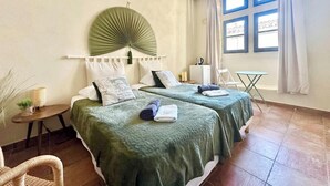 A hotel room with two beds, a small table, a chair, a hanging light fixture, and a window with curtains. - Maison Cosy Lodge, 15 p, clim, wifi, cour, garage (Montfrin)