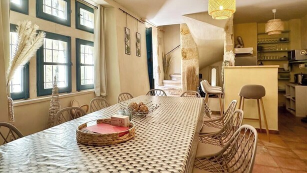 A dining area with a long table, chairs, and a kitchen in the background. - Maison Cosy Lodge, 15 p, clim, wifi, cour, garage (Montfrin)