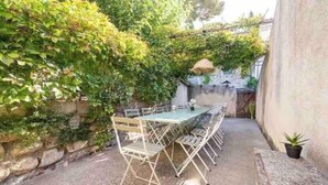 An outdoor patio with a table and chairs, surrounded by greenery and a stone wall. - Maison Cosy Lodge, 15 p, clim, wifi, cour, garage (Montfrin)