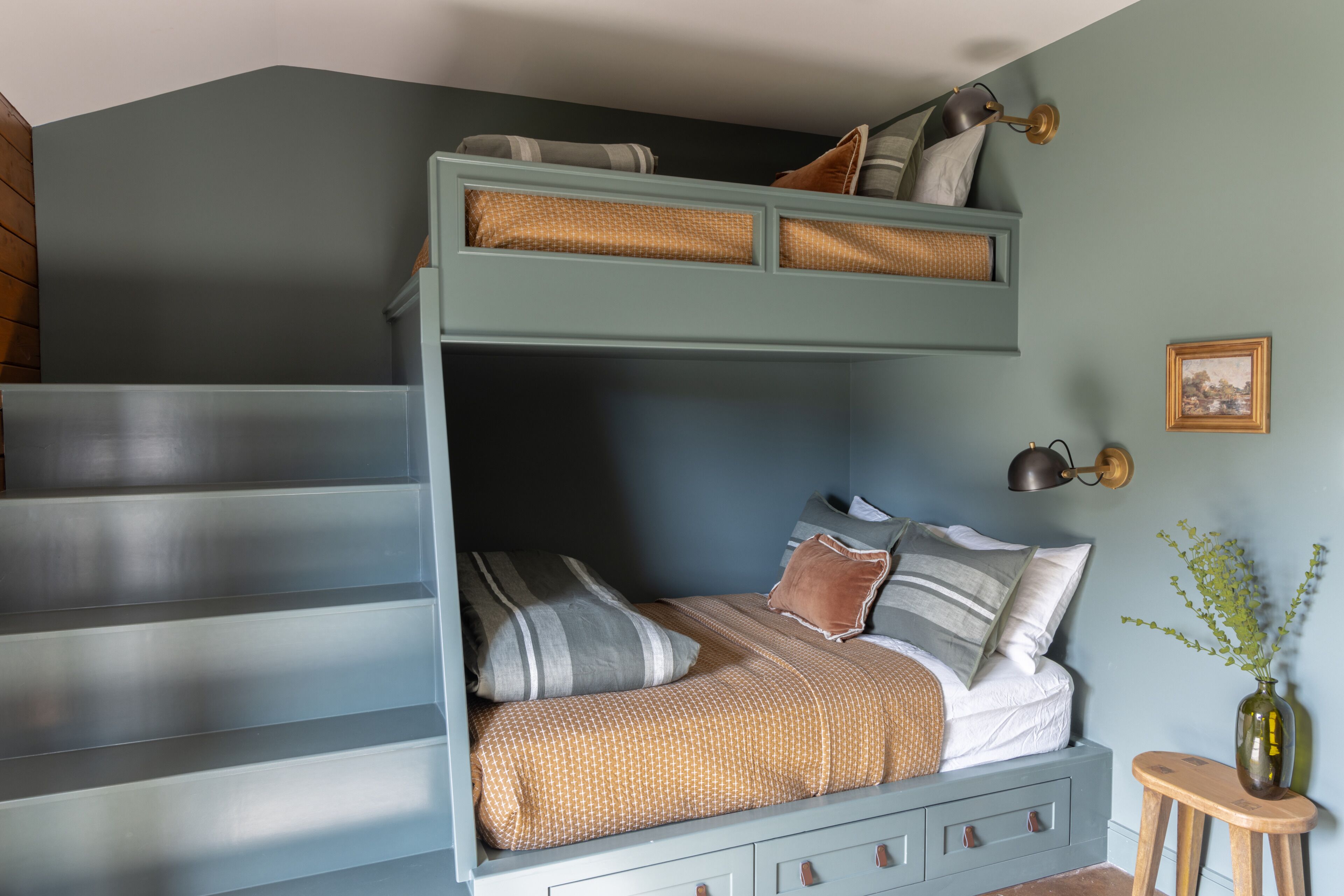 A bunk bed with a lower bunk, a bedside table, a lamp, and a vase with plants.